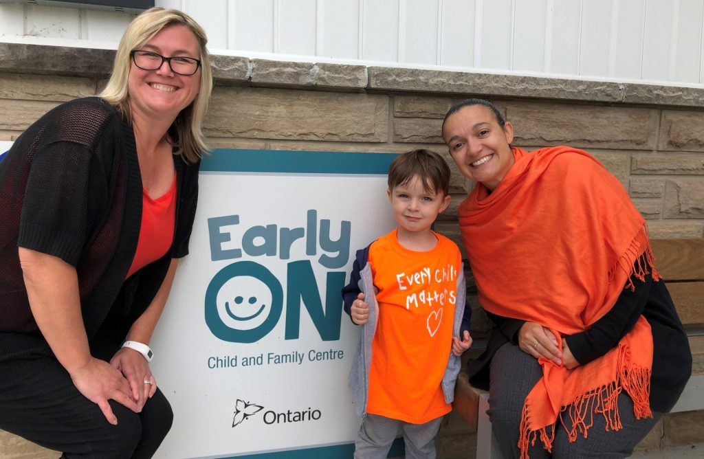 Two workers and a kid are posing near the sign of Early Years Centre Ontario