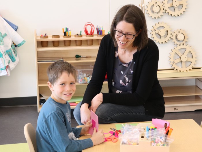 A kindergarten teacher is sitting near a boy who is doing a craft  in EarlyON Years Centre